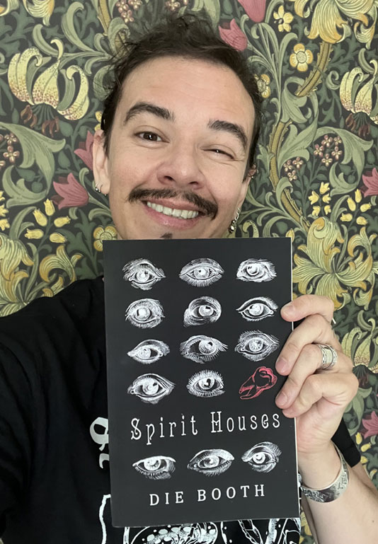 Author Die Booth smiling holding a copy of Spirit Houses. He is a white middle aged man with dark hair and moustache, wearing a black tshirt. The book cover is black, with the title and line drawings of eyes in white, and a single line drawing of a tooth in red.