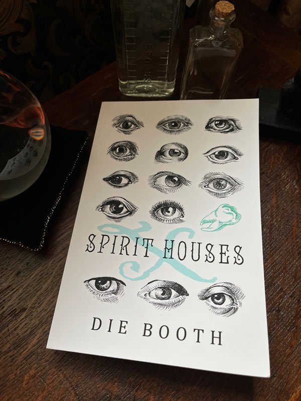 Image shows Spirit Houses X by Die Booth, a white book with black line drawings of eyes and a teal green X and line drawing of a tooth, set on a table with a crystal ball and some old glass bottles
