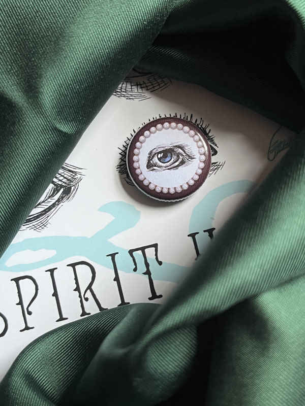 Image shows the cover of Spirit Houses X mostly covered in green silk. You can just about see a white background, black text and a green swirl, with a badge showing an illustrated blue eye resting on top of the book cover.