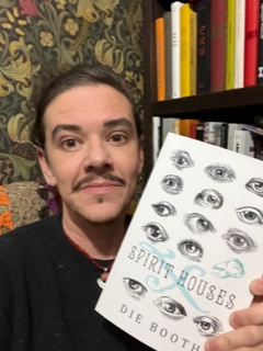 Photo of author Die Booth (white man with dark eyes, hair and moustache) holding a copy of Spirit Houses (white cover with black ink eyes and one green ink tooth, with title in black and a swirly green 'X' behind the title)